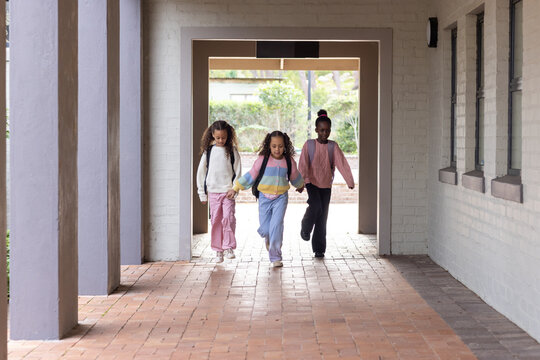 Three schoolgirls walking toward camera in covered brick corridor carrying backpacks and smiling
