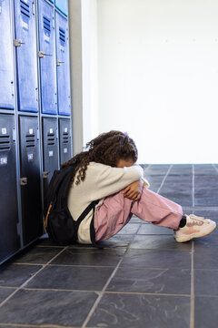 African adolescent female student sitting against blue lockers on tiled floor, with black backpack