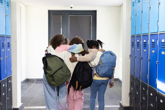 African American teen females walking arm-in-arm in school hall by blue lockers carrying backpacks