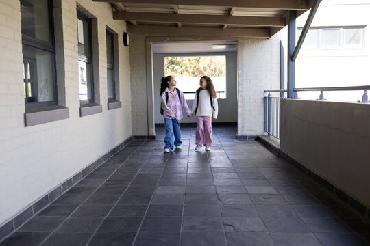 Two African American female youths walking hand in hand in covered school corridor with backpacks