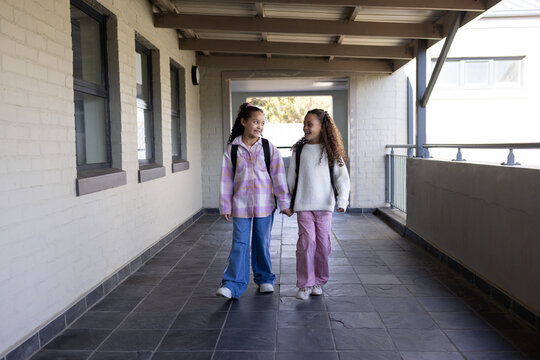 Two preteen African American girls walking along school corridor holding hands, carrying backpacks