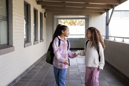 African American youth female friends holding hands and walking in school corridor with backpacks