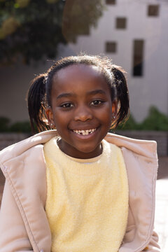 African schoolgirl smiling, standing in sunlit courtyard wearing beige coat yellow sweater pigtails