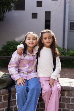 African American school-age girls sitting close together on brick ledge in courtyard with backpacks