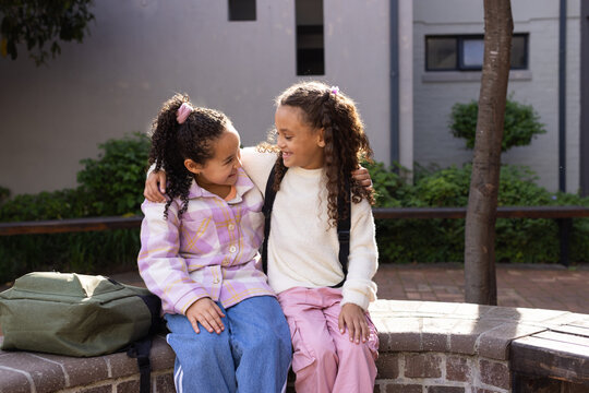 African American schoolgirls sitting and hugging on curved brick bench in courtyard, green backpack