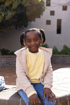 African American schoolgirl sitting on bench in courtyard wearing hooded coat and backpack