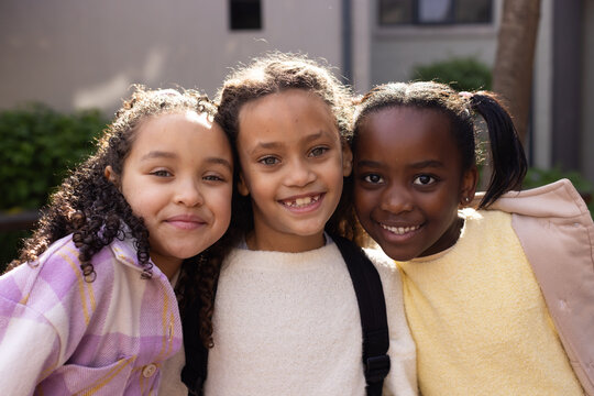3 African American schoolgirls standing in sunny courtyard wearing sweaters backpack straps showing