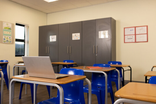 Silver laptop is sitting on front wooden desk in classroom with blue chairs and gray cabinets