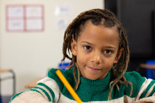 School-age African American boy wearing green sweater holding yellow pencil at classroom desk