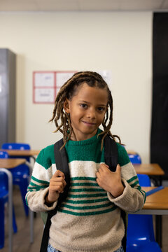 African American schoolboy standing at classroom front holding black backpack in striped sweater