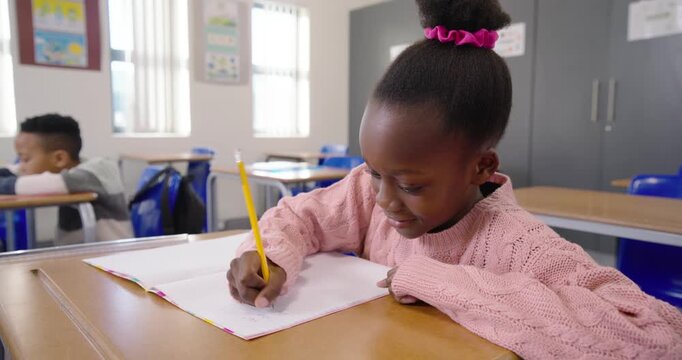 Child girl in pink sweater sitting at school desk, writing classwork in notebook with yellow pencil