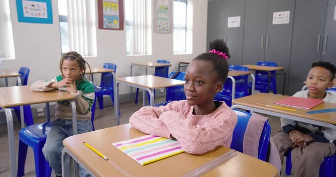 Schoolchildren watching front girl smiling replying to teacher prompt at desk with striped notebook