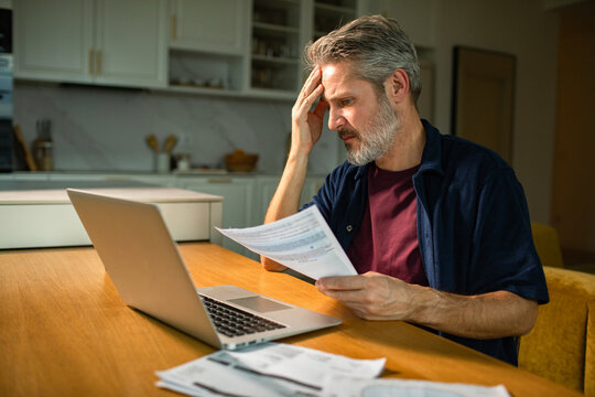 Middle-aged man stressed over bills on laptop at home kitchen