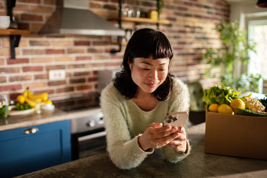 Young woman using smartphone in home kitchen with fresh groceries