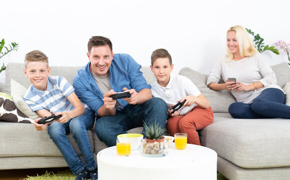 Father and his two young sons sit on the sofa holding game controllers, smiling and excitedly playing a console video game together. In back, mother sits relaxed on the couch using smartphone.