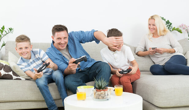 Father and his two young sons sit on the sofa holding game controllers, smiling and excitedly playing a console video game together. In back, mother sits relaxed on the couch using smartphone.