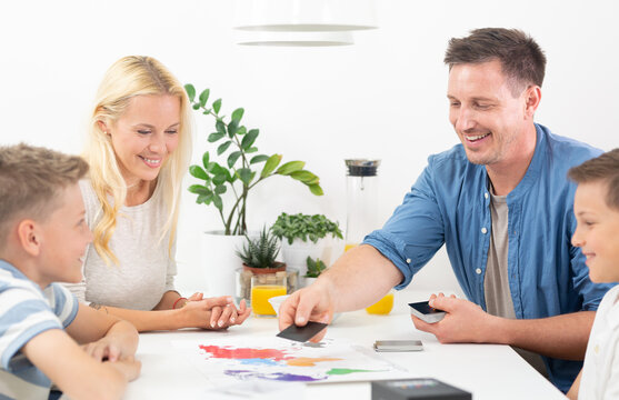 Happy family playing a world map educational board game together around table. Father placing card on world map board game while family watches and smiles.