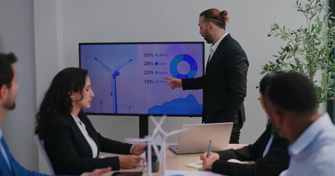 Middle Eastern executive points at renewable energy chart on display during corporate team meeting. Male speaker explains turbine metrics and answers questions