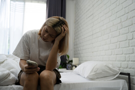 Sad teenage girl receives bad news online on her smartphone, sitting on her bed in her bedroom at home.
