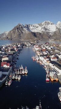 Aerial view of Henningsvaer fishingg village with boats and traditional houses against snowy mountains in wintere, Lofoten Islands, Norway