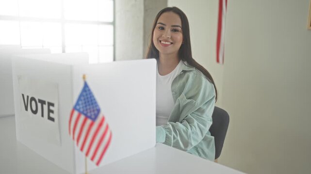Young hispanic woman smiling at a voting booth holding a ballot with an american flag visible in a polling building; civic pride.
