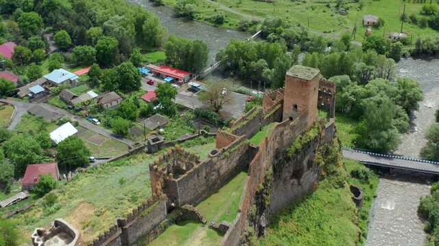 Khertvisi Fortress dominates valley, aerial view beside river channels, village homes and a bridge, supporting defensive architecture, regional geography, heritage tourism and Georgian history.