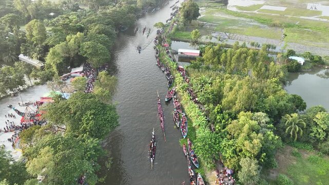 Gopalganj, Bangladesh - 30 October 2023: Aerial view of a traditional boat race on a narrow canal with many long wooden boats lined up along the lush green banks.