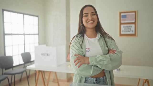 Woman smiling with arms crossed, voted sticker visible beside ballot box in polling building; pride civic duty.