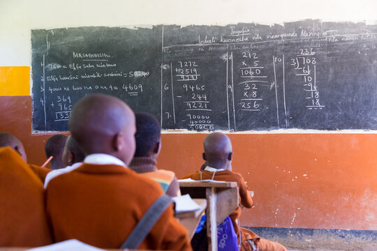 African elementary school students in uniforms sitting at wooden desks, looking at handwritten math equations and Swahili text written in chalk across a large blackboard in a bright classroom.