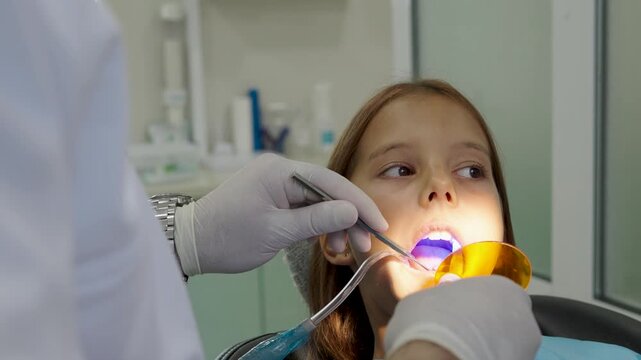 Pediatric dentist using UV curing light lamp on tooth filling for young girl.
