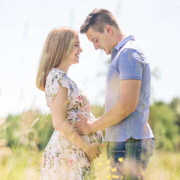 Side view of a happy pregnant couple embracing gently and holding the belly in a sunlit grassy field during summer. Romantic maternity concept of love, family preparation, and new life.