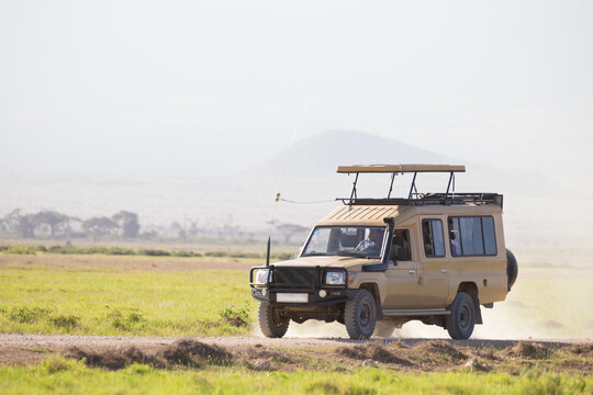A beige 4x4 safari truck with a raised pop-up roof driving along a dirt road through the open grassy savanna plains of an African national park during a game drive excursion.