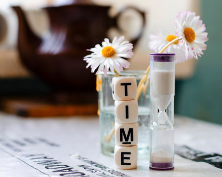 Time inscription made up of small cubes, hourglass and decorative white flowers in a glass in the background, selective focus, blurred background