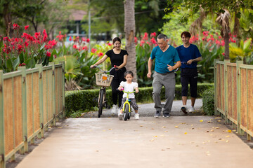 Obraz premium Joyful Asian young girl while riding a bicycle with her grandfather in park on sunny day. girl kid on bicycles having fun in city park. outdoor activitie with daughter father mother and grandfather