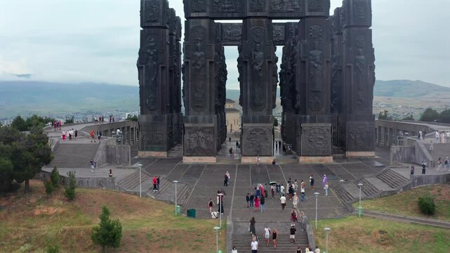Aerial drone flies forward toward the Chronicle of Georgia monument in Tbilisi, approaching monumental columns and visitors for landmark tourism, Georgian history, architecture, destination promotion