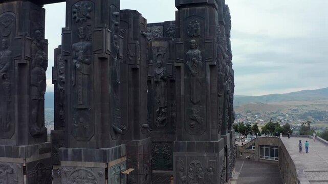 Aerial drone pulls backward beside the Chronicle of Georgia monument in Tbilisi, revealing carved columns, religious reliefs and city views for cultural tourism, architecture and history documentaries