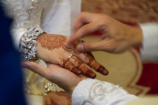 Close-up of a groom placing a golden wedding ring on the bride's hand, decorated with intricate henna (mehndi) designs and a diamond bracelet during a traditional wedding ceremony.
