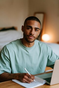 Young man looking at camera while writing in notebook beside laptop in bedroom for online study and remote work background