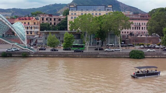 Tour boat crosses river-level aerial view beside Rike Park and the Bridge of Peace in central Tbilisi, supporting river tourism, sightseeing, urban transport and Georgian destination promotion.