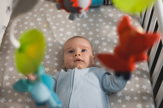Cute infant baby lying in crib and looking up at hanging colorful mobile toys in a cozy nursery.