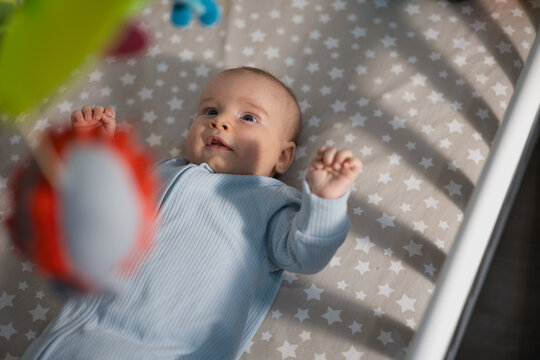 Side view of cute infant baby lying in crib and looking up at hanging colorful mobile toys in a cozy nursery.