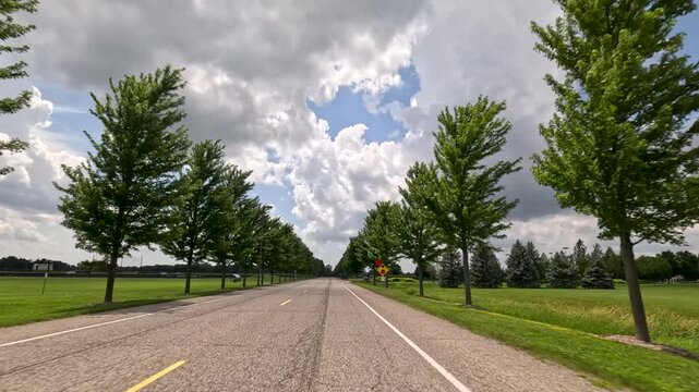 Moving vehicle perspective along a straight tree-lined college campus road near outdoor sports fields and signage. Captured at University of Illinois Springfield.
