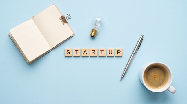 Workspace with an open notebook, lightbulb, pen, coffee cup, and scrabble tiles spelling "STARTUP" on a light blue background.