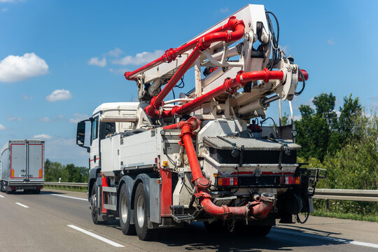 Concrete pump truck travels along highway with folded red boom and hydraulic pipes. Heavy construction vehicle represents mobile machinery, infrastructure logistics and industrial transport