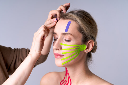 Young woman with tapes on her face and hands specialist measuring face contour for cosmetic procedures, skin care and aesthetic correction concept, light background.
