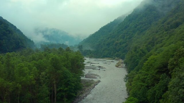 Drone flies forward above the Aragvi River in Georgia, following a broad mountain channel through dense forest and misty slopes for nature tourism, conservation and destination advertising campaigns.