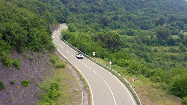 Aerial drone flies forward above a mountain highway near Zhinvali Reservoir in Georgia, following a car through dense forest for scenic driving, road tourism, transport media and travel campaigns.