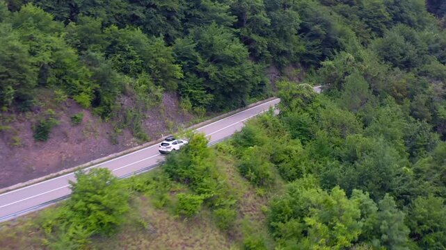Aerial drone moves sideways above a forest highway near Zhinvali Reservoir in Georgia, tracing a car past a cut slope for transport planning, road infrastructure, tourism access and travel media.