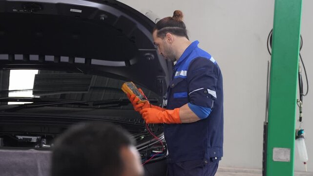 Mechanic and technician in workshop performing car hood diagnostic with multimeter, maintenance and repair service checking engine electrical system, glove use, testing battery with meter
