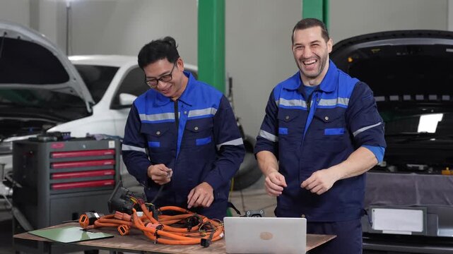 Mechanic technician garage car repair with diagnostic laptop, wiring harness voltage cable testing on electric vehicle workbench, teamwork of two male workers smiling during maintenance
